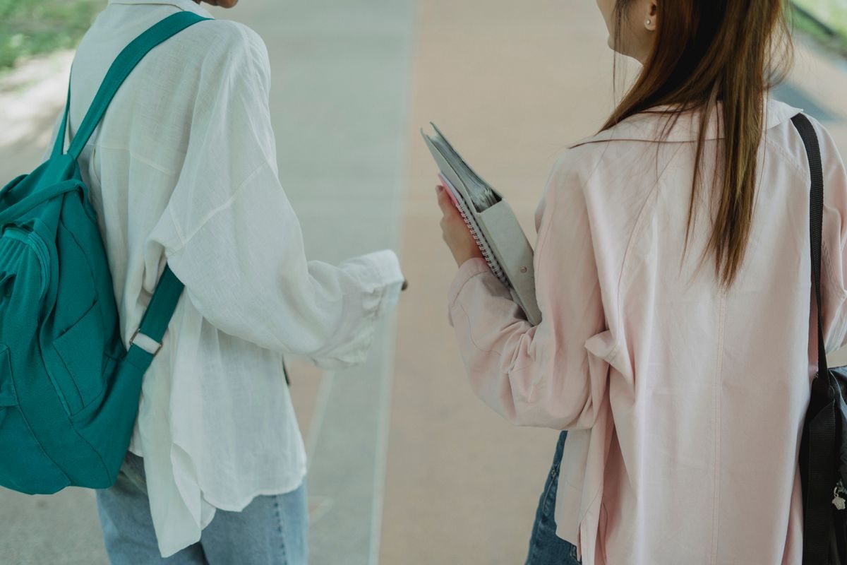 歩道を並んで歩く2人の学生の後ろ姿。リュックを背負い、ノートを抱えてキャンパスへ向かうイメージ