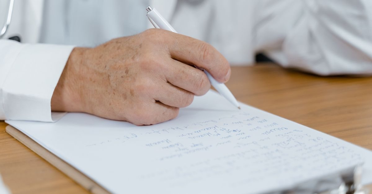 白衣の医師がクリップボードの書類にペンで記入している手元。有期再認定の診断書は所定の様式で主治医に依頼する