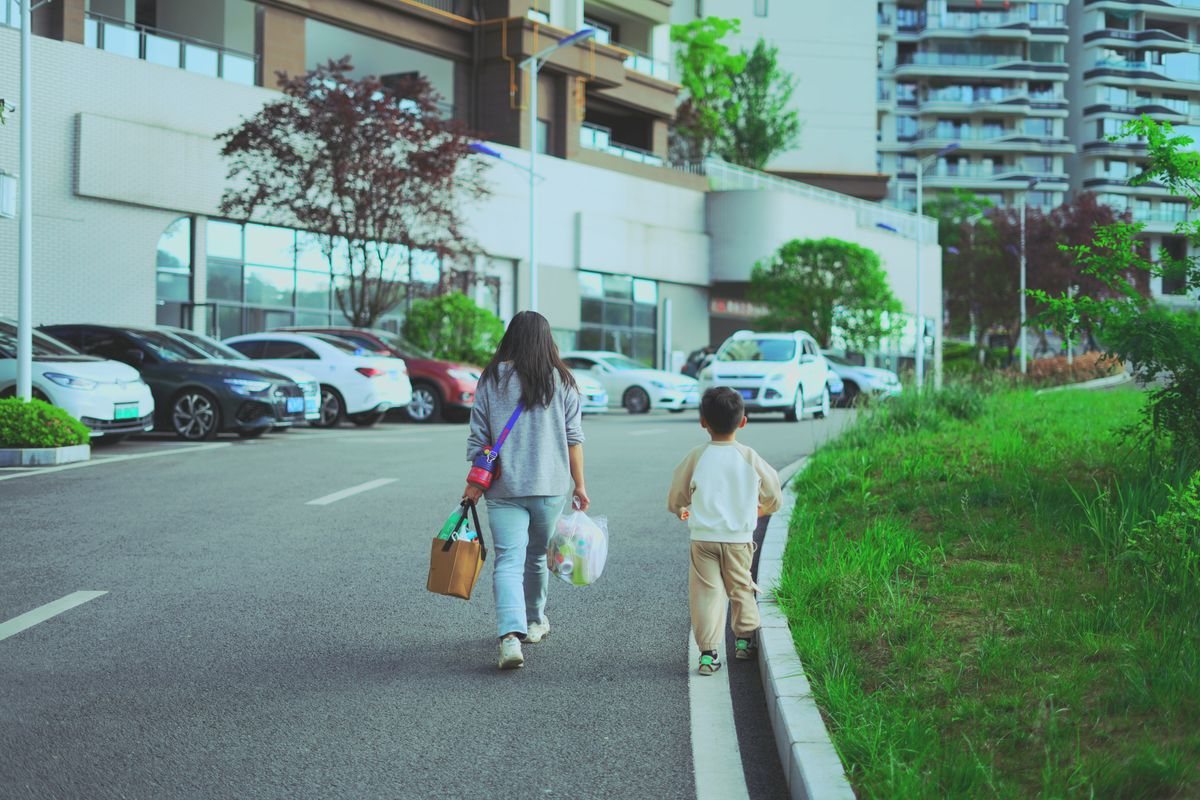 マンションの並ぶ道を、荷物を持った大人と小さな子どもが歩いて帰っていく後ろ姿。園のお迎えの帰り道のイメージ