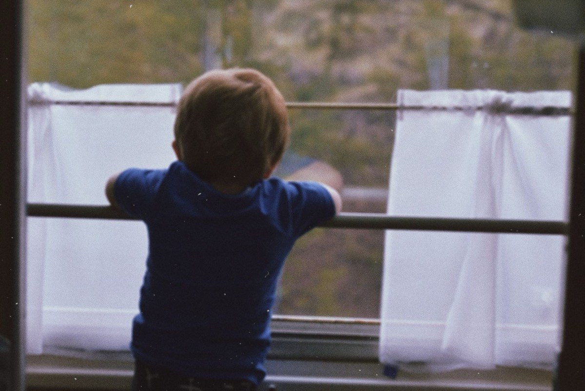 白いカーテンの窓辺で外を静かに見ている子どもの後ろ姿。園や学校で声が出にくい子のイメージ