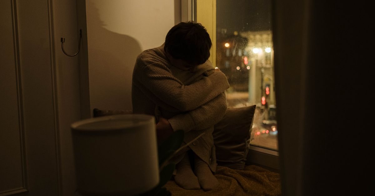 夜、明かりを落とした部屋の窓辺でひざを抱えてうつむく大人。子どもの診断をまだ受け止めきれずにいる夜のイメージ