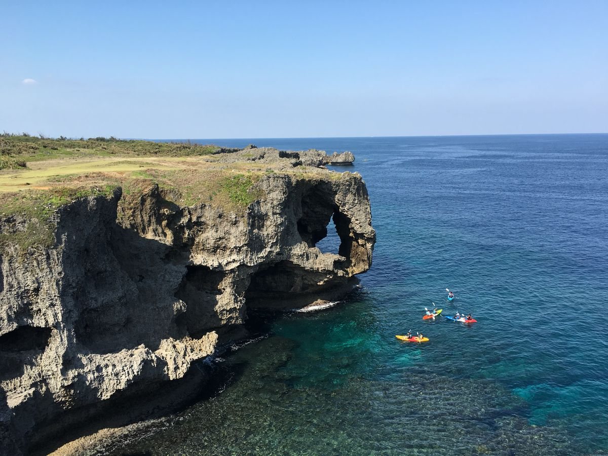 沖縄本島の万座毛の崖と、どこまでも続く青い海。遠くに小さなカヤックが浮かぶ。2年次に沖縄伊計本校へ向かうイメージ