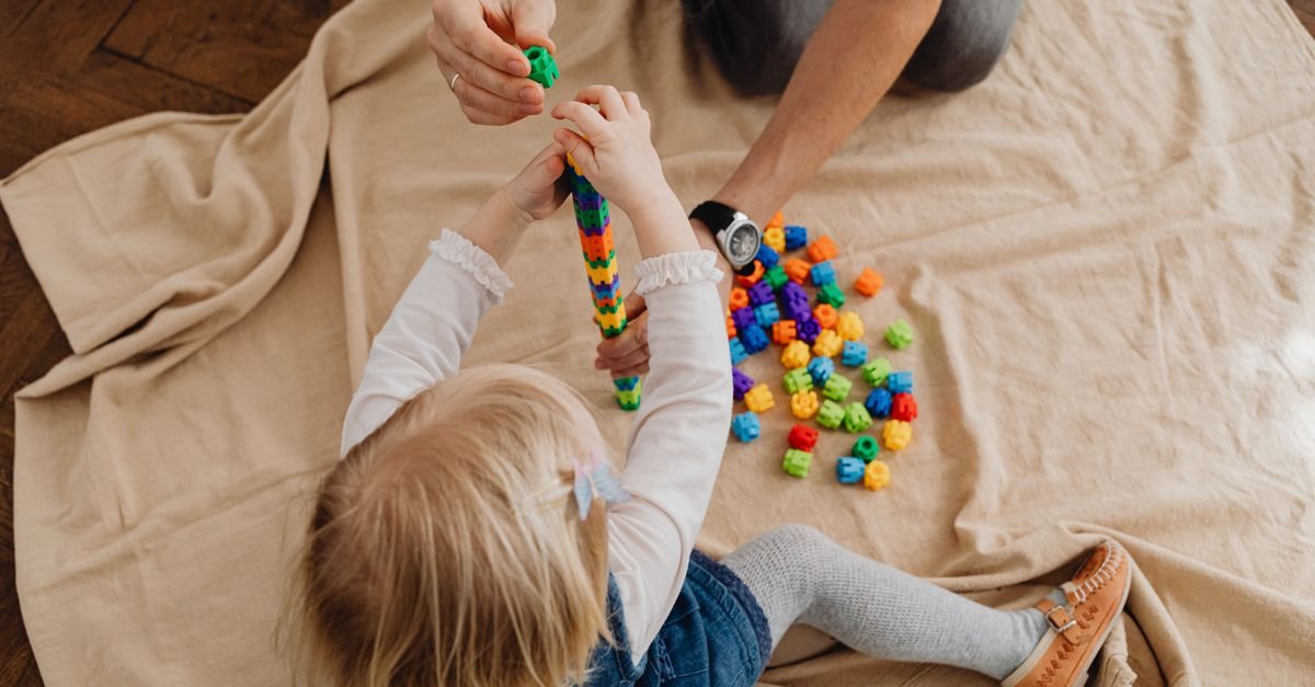 布の上でカラフルなブロックをつなげて遊ぶ幼児と、向かいで手を添える大人（親子教室の親子あそびのイメージ）