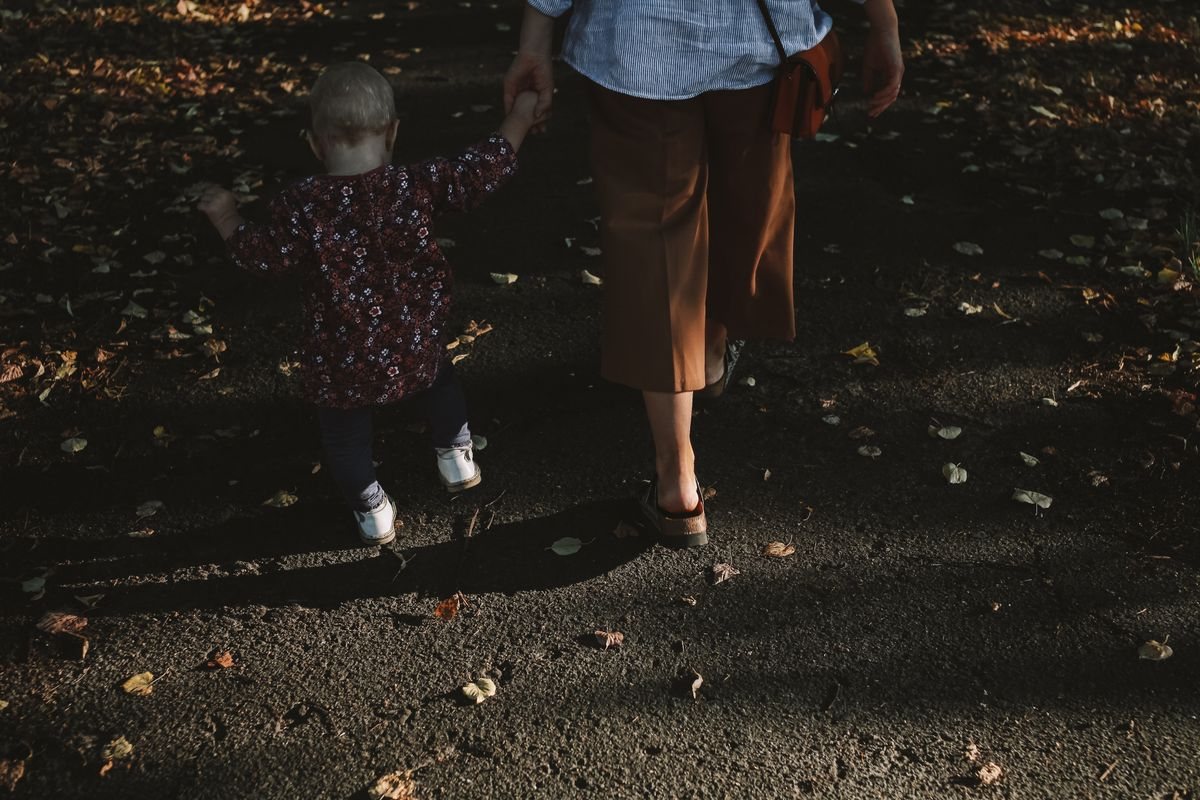 落ち葉の道を大人と手をつないで歩く幼児の後ろ姿。親子教室へ向かう道のイメージ
