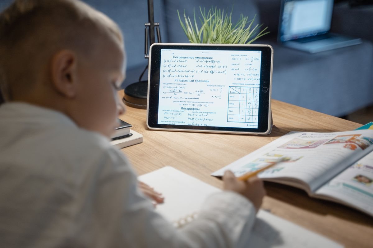 机の上のタブレットと教科書を前に、ノートに書き込む子どもの後ろ姿。読み上げや画面の作りが合うかを、家で実際に試してみる場面のイメージ