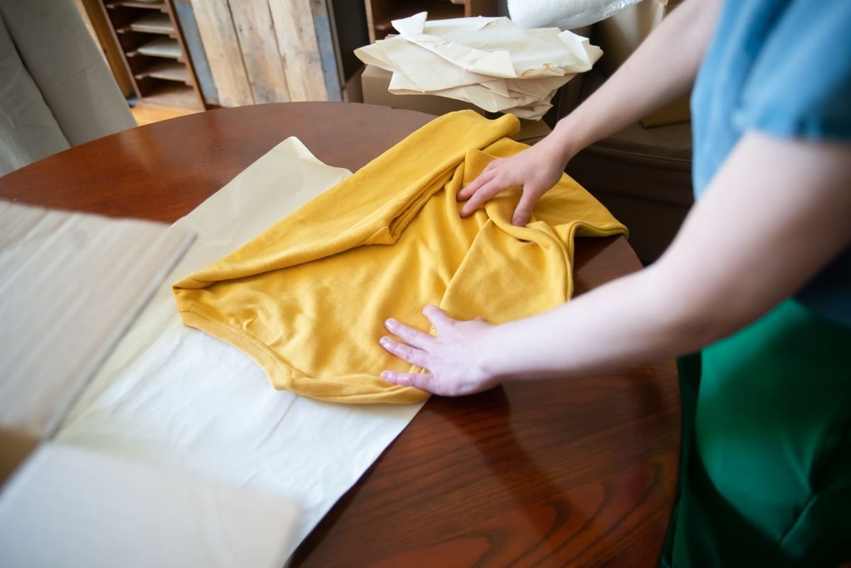 木のテーブルに黄色いTシャツを広げ、両手でしわを伸ばしている手元。服の前と後ろ、表と裏を目で確かめる場面のイメージ